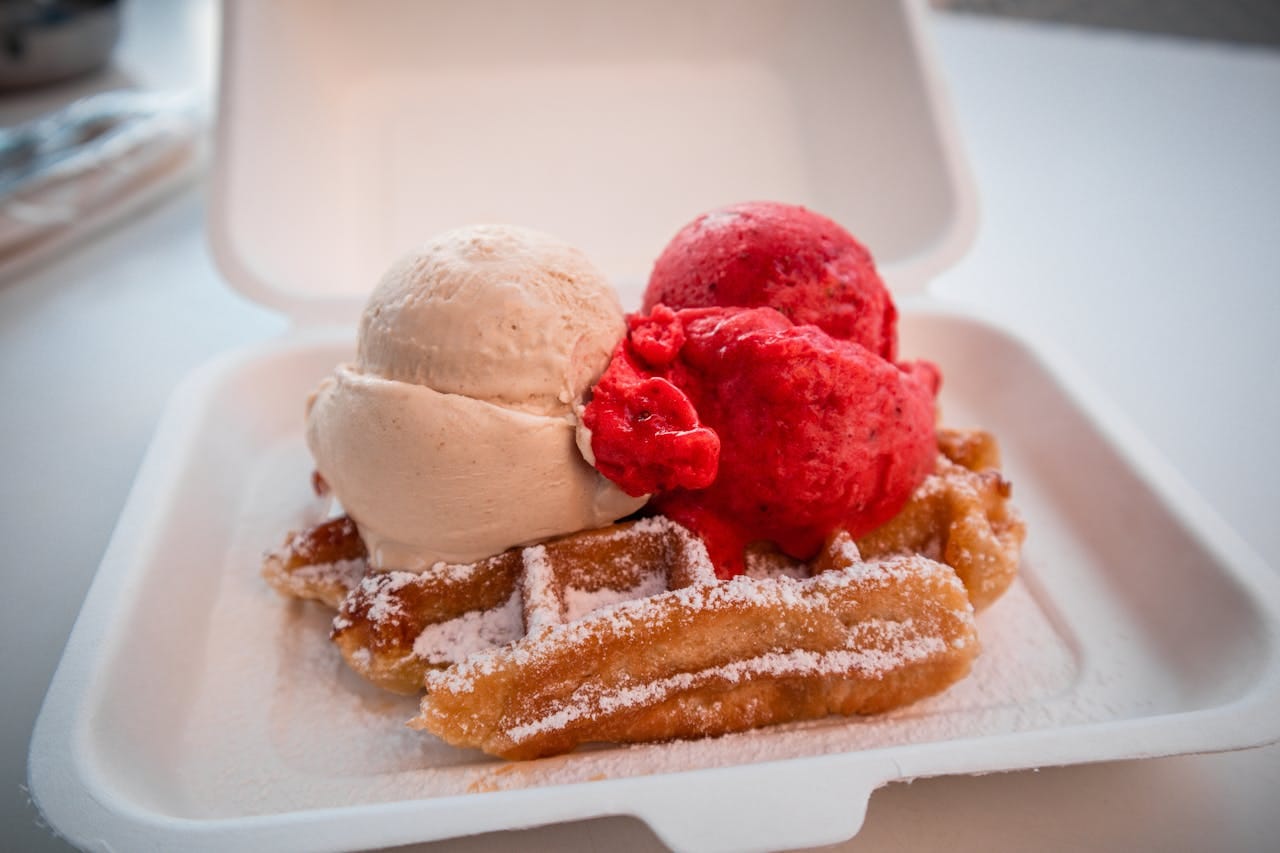 Close-up of a tasty waffle topped with vanilla and strawberry ice cream in a takeout container.